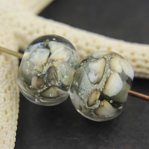 May include: Two clear glass beads with a mix of white, gray, and brown inclusions. The beads are strung on a copper wire, with a starfish in the background. The beads have a unique, organic appearance.