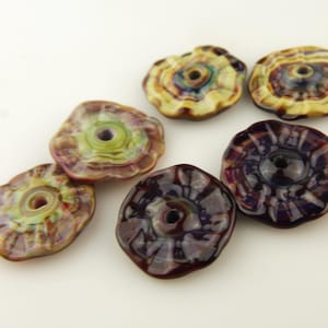 Könnte beinhalten: Sieben handgefertigte Glasperlen in Blumenform. Die Perlen haben ein Loch in der Mitte und sind in Braun-, Lila-, Grün- und Gelbtönen gehalten. Die Perlen haben eine glänzende Oberfläche und sind auf einer weißen Oberfläche angeordnet.