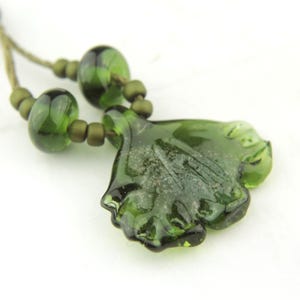 May include: A dark green glass pendant shaped like a ginkgo leaf, suspended on a cord with two matching translucent green glass beads and smaller matte olive green spacer beads on each side of the central leaf-shaped glass element.