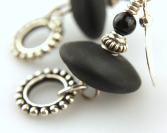May include: Close-up of black stone earrings with silver accents. The earrings feature smooth, oval black stones, small black beads, and decorative silver metalwork. The design includes a hook closure.