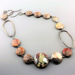 May include: A handcrafted necklace featuring a mix of round, textured beads in shades of brown, orange, and cream, interspersed with silver-toned metal links. The focal bead is a larger, colorful, patterned bead. The necklace has a hook clasp.