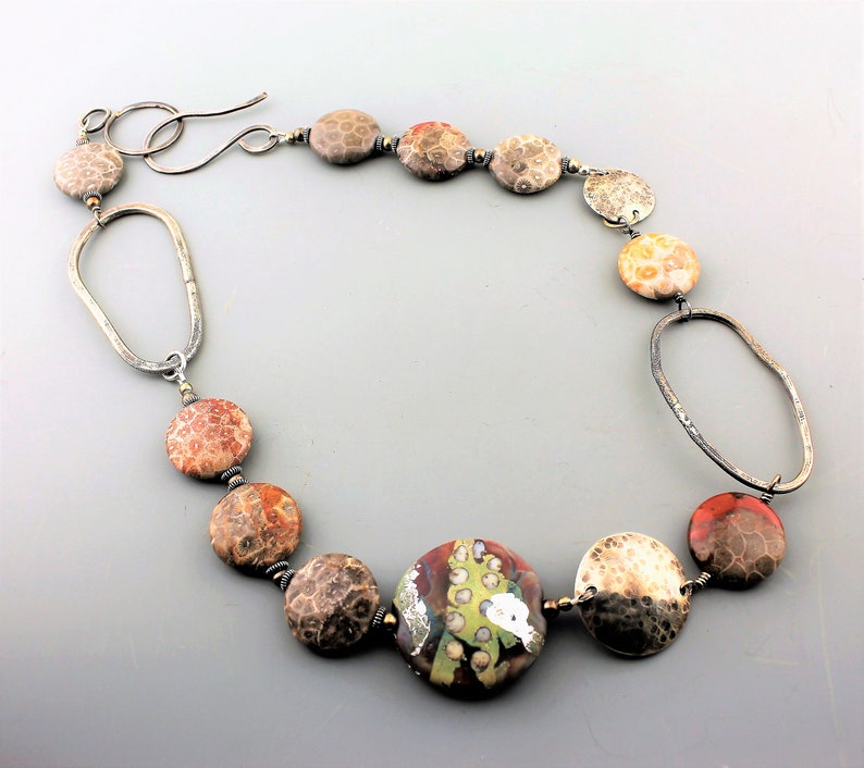 May include: A handcrafted necklace featuring a mix of round, textured beads in shades of brown, orange, and cream, interspersed with silver-toned metal links. The focal bead is a larger, colorful, patterned bead. The necklace has a hook clasp.