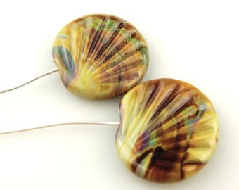 Könnte beinhalten: Zwei handgefertigte Glasperlen mit muschelartigem Design. Die Perlen sind hauptsächlich gelb und braun, mit grünen und violetten Streifen. Jede Perle hat ein Loch zum Auffädeln. Auf weißem Hintergrund.