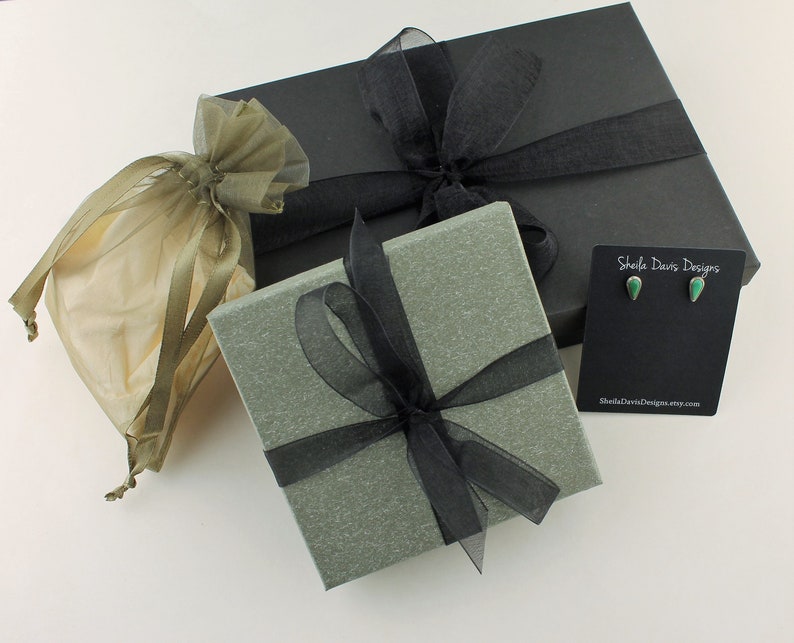 May include: Gift boxes and jewelry packaging. A black box and a gray box are tied with black ribbon. A small pair of green teardrop earrings on a black card. A small beige organza gift bag.