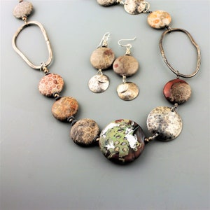 May include: A handcrafted necklace and earring set featuring large, round, and oval beads in shades of brown, red, and gray. The necklace includes a large, round, green and brown focal bead. The earrings have silver accents.