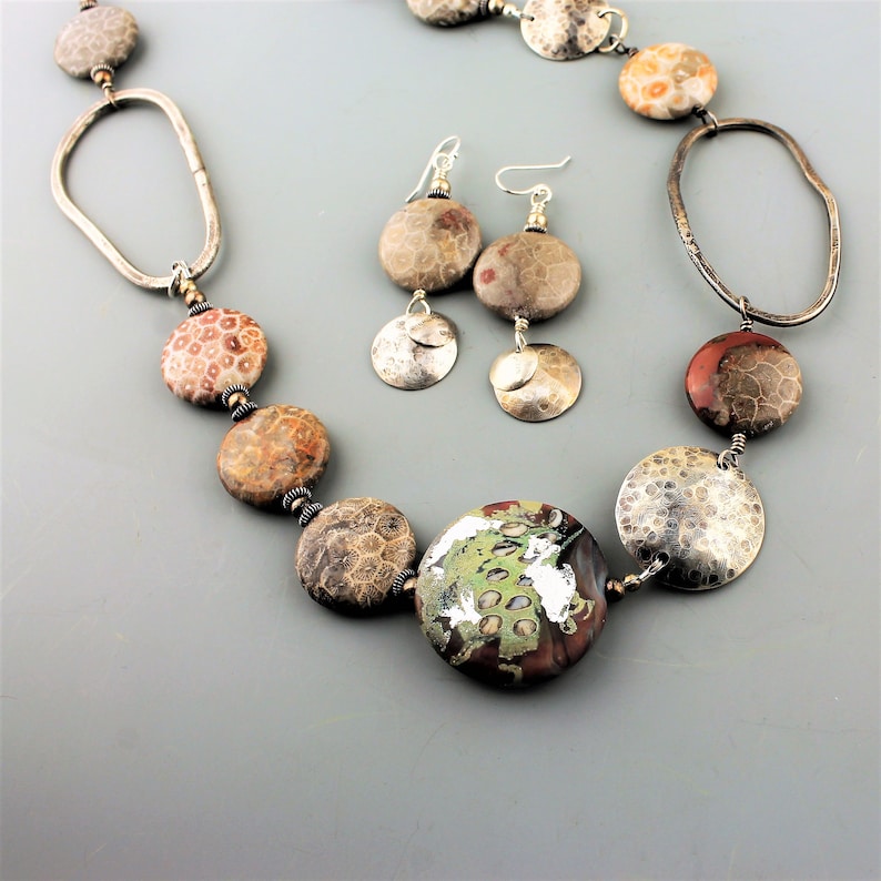 May include: A handcrafted necklace and earring set featuring large, round, and oval beads in shades of brown, red, and gray. The necklace includes a large, round, green and brown focal bead. The earrings have silver accents.