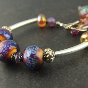 May include: A silver bangle bracelet with purple and orange glass beads. The beads have a floral pattern and iridescent finish.