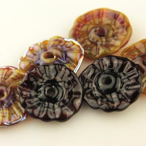 Könnte beinhalten: Sechs handgefertigte Glasperlen in Blütenform mit einem zentralen Loch und einem gerüschten Rand. Die Farben umfassen Bernstein, Braun, Lila und Schwarz. Diese Perlen eignen sich für die Schmuckherstellung und andere Bastelprojekte.