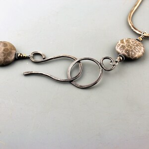 May include: Close-up of a silver necklace with a unique clasp. The necklace features two large, round, fossilized coral beads in shades of gray and brown. The clasp is made of hammered silver, with a hook and loop design.