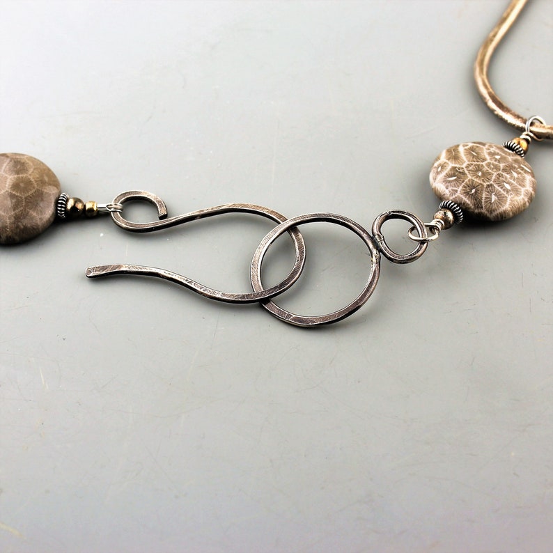 May include: Close-up of a silver necklace with a unique clasp. The necklace features two large, round, fossilized coral beads in shades of gray and brown. The clasp is made of hammered silver, with a hook and loop design.
