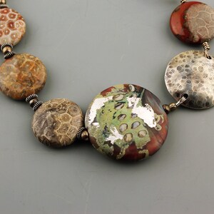May include: A necklace featuring a variety of round beads in shades of brown, red, green, and silver. The beads have unique textures and patterns, including a fossil-like design and hammered metal discs. The necklace is strung together with small, silver-colored connectors.