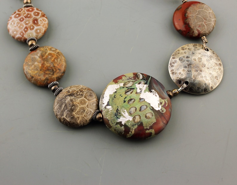 May include: A necklace featuring a variety of round beads in shades of brown, red, green, and silver. The beads have unique textures and patterns, including a fossil-like design and hammered metal discs. The necklace is strung together with small, silver-colored connectors.