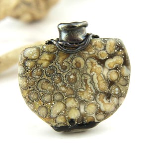 May include: A pendant featuring a polished, rounded stone with a complex pattern of beige, tan, and dark brown circular inclusions. The stone is topped with a dark, metallic, textured cap that wraps around the upper edge of the piece.