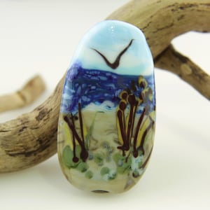 Puede incluir: Una cuenta de vidrio artesanal con una escena de playa. La cuenta presenta un cielo azul con un pájaro volando, un océano azul oscuro y una playa arenosa con plantas marrones y amarillas. La cuenta mide aproximadamente 3,8 cm de largo.
