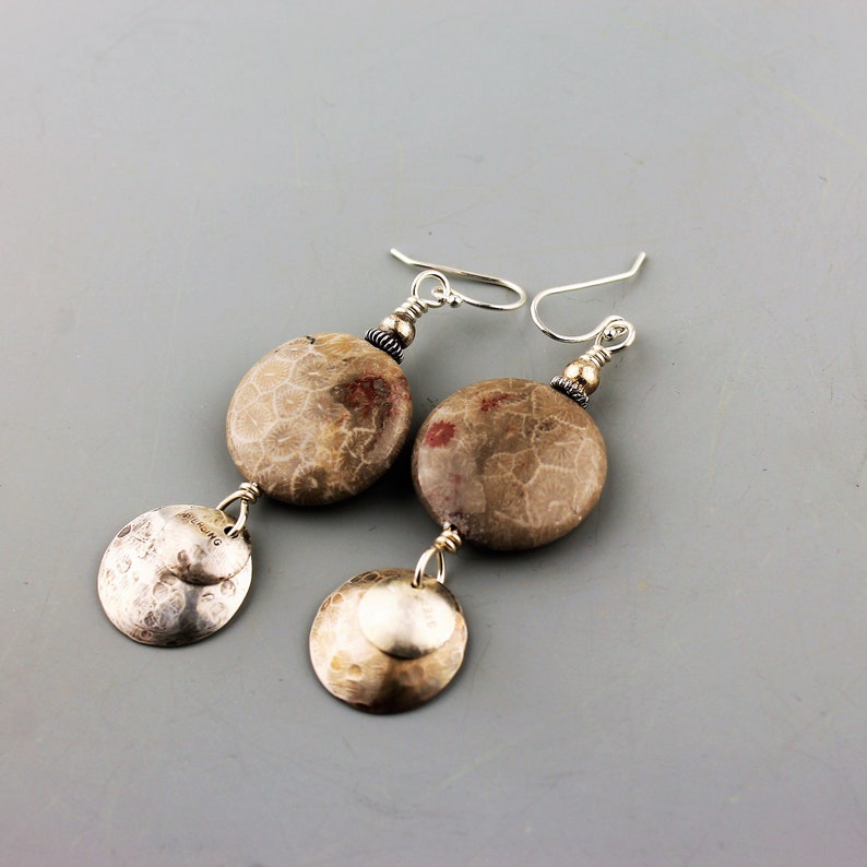 May include: A pair of dangle earrings featuring fossilized stone beads and hammered silver discs. The earrings have a neutral color palette with silver hooks and accents. The fossilized stone beads have a beige and brown color with a unique pattern.