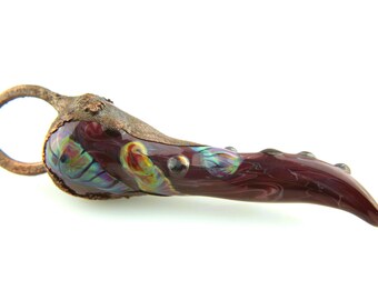 May include: A curved, horn-shaped pendant featuring a deep burgundy glass body with iridescent swirls of blue, green, and yellow. The piece is capped with a textured, weathered copper-toned metal fitting and includes a circular metal bail.
