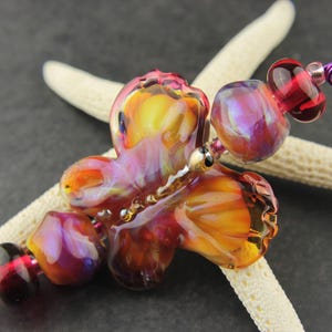 Op de afbeelding: Een kleurrijke glazen vlinderkraal met rode, oranje, gele en paarse swirls. De kraal is bevestigd aan een witte zeester met een paarse en gouden draad.