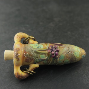 May include: A small, hand-blown glass pendant in the shape of a vase with a gold-colored metallic finish. The vase has a textured surface with a green, brown, and gold color scheme. The vase has a cluster of purple glass beads on the side. The vase has two gold-colored metal loops at the top.