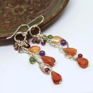 Może przedstawiać: Wiszące kolczyki ze srebrnymi haczykami i łańcuszkami. Kolczyki mają różnorodne kolorowe kamienie szlachetne, w tym pomarańczowe, fioletowe, zielone i czerwone koraliki. Kamienie szlachetne są ułożone kaskadowo, tworząc unikalny dodatek.
