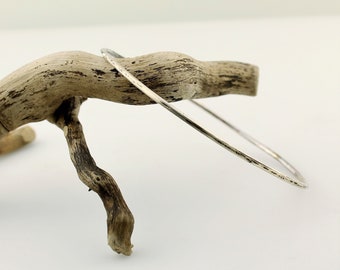 May include: A thin silver bangle bracelet with a hammered finish. The bracelet is displayed on a piece of driftwood.