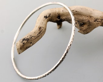 May include: A silver bangle bracelet with a hammered texture. The bracelet is displayed on a piece of driftwood.