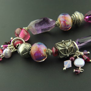 May include: A purple and pink beaded necklace with silver accents. The necklace features a variety of beads, including amethyst, glass, and metal. The necklace is accented with small silver charms and a silver pendant.