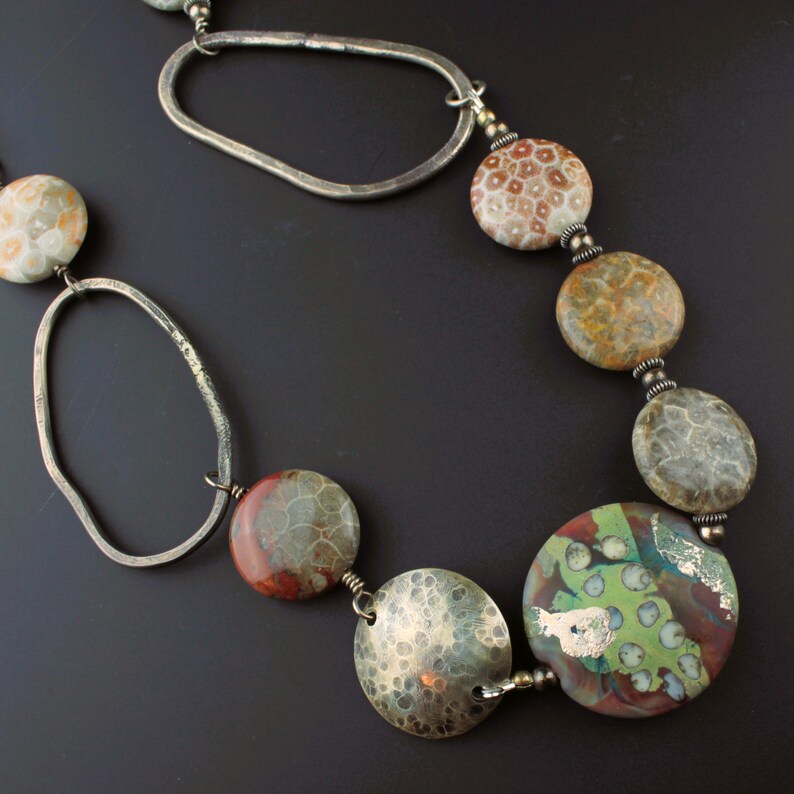 May include: A handcrafted necklace featuring large, round, and oval beads in shades of brown, red, green, and silver. The necklace includes hammered silver disc beads and irregular silver links, creating a unique and artistic design.