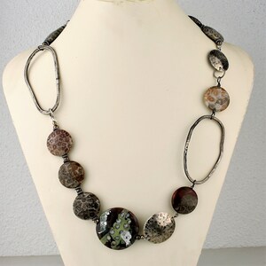 May include: A handcrafted necklace featuring a mix of textures and colors. The necklace includes large, round, patterned beads in shades of brown, gray, and tan, interspersed with hammered silver-tone metal elements and a large, green and brown focal bead.