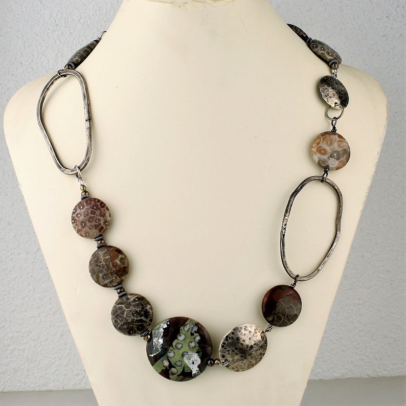 May include: A handcrafted necklace featuring a mix of textures and colors. The necklace includes large, round, patterned beads in shades of brown, gray, and tan, interspersed with hammered silver-tone metal elements and a large, green and brown focal bead.