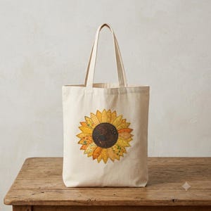 May include: A cream-coloured canvas tote bag featuring a large, central sunflower graphic. The sunflower is composed of various yellow, orange, and gold fabric patches with intricate stitching, showcasing a dark brown centre with a textured pattern.