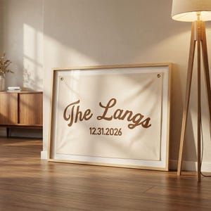 Puede incluir: Impresión en lienzo enmarcada con el texto "The Langs" y la fecha "12.31.2026" en marrón. El lienzo está dentro de un marco de madera clara, apoyado contra una pared. El diseño tiene un estilo minimalista y moderno.