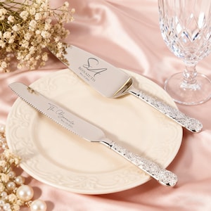 Può includere: Set di coltello e paletta per torta argentati con manici decorati, su un piatto bianco. Il server è inciso con "SA BONAIUTO May 19 2023", e il coltello con "The Bonaiutos May 19 2023". Il set è esposto con fiori e un bicchiere di cristallo.