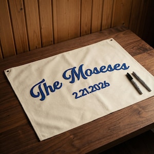 Puede incluir: Una pancarta crema con el texto bordado "The Moseses" en azul y la fecha "2.21.2026". Dos bolígrafos descansan sobre la pancarta, colocada sobre una superficie de madera.