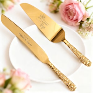 Può includere: Set di coltello e paletta per torta dorati con dettagli floreali sul manico. I nomi "Ethan & Sophia" e la data "9.16.2023" sono incisi. Il set è presentato su un piatto bianco, con fiori rosa sullo sfondo.