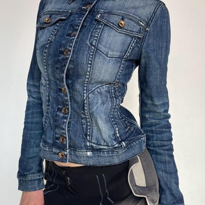 May include: A fitted, dark-wash denim jacket with a collared neckline and long sleeves. The jacket features button closures, chest pockets, and a tailored silhouette. The denim has a faded, worn appearance, and the buttons are a brass colour.