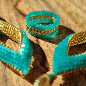 Könnte beinhalten: Ein Set aus türkisfarbenen und goldenen Perlen-Schmuck mit einem Ring und einem Paar Ohrringen. Der Ring und die Ohrringe sind aus kleinen Perlen gefertigt, die in einem geometrischen Muster miteinander verwoben sind.