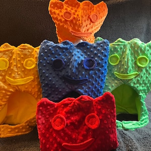 May include: Five plush toy headbands in different colours, each with a smiling face. The headbands are orange, blue, green, red, and yellow.