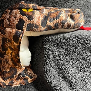 May include: A brown and black snake costume with a yellow eye and a red tongue. The costume is made of fabric and has a hood.