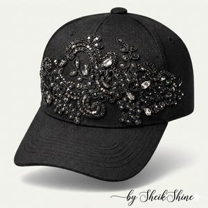 May include: Black baseball cap with ornate, dark-colored beading and crystal embellishments. The cap features a curved brim and a button on top. The text "by Sheik Shine" is at the bottom.