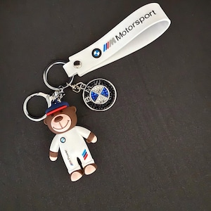 May include: A white keychain with a blue and white BMW Motorsport logo and a brown bear wearing a white racing suit with the BMW logo. The bear is holding a key ring with a black and blue BMW logo with rhinestones.