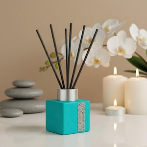 May include: A teal reed diffuser with black reeds and a silver collar. A vertical strip of small, silver embellishments decorates the front. White candles and a stack of grey stones are in the background. White orchid flowers are also visible.