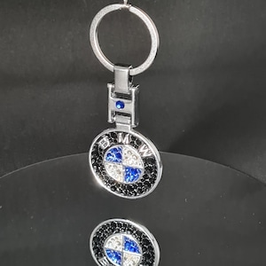 Pode incluir: Um chaveiro prateado com um logotipo BMW preto e branco. O logotipo é feito de strass e tem um centro azul.