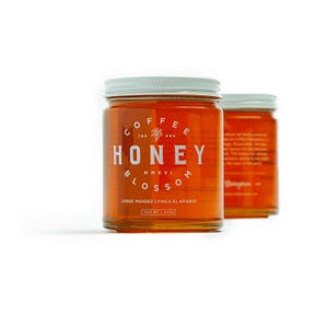 May include: Two glass jars of honey with white lids. The front jar has a label that reads "Coffee Honey Blossom MMXVI Jorge Mendez | Finca El Apiario 12 OZ WT | 340G".