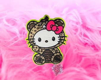 May include: A character figure featuring a white cat face with black eyes and whiskers, wearing a brown and black leopard print hooded onesie with a bright pink glittery bow on the side, outlined in a vibrant neon yellow border.