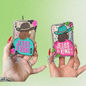 May include: Two air fresheners shaped like playing cards, each featuring an illustration of a figure wearing a hat and the text "JESUS IS KING." One card has a brown hat and teal accents, the other has a teal hat and pink accents.