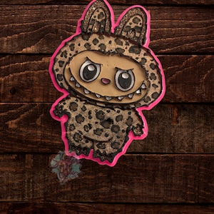 Puede incluir: Una criatura caprichosa y caricaturesca con un disfraz de estampado de leopardo con un contorno rosa. La criatura tiene ojos grandes, una sonrisa con dientes y orejas de conejo. El fondo es una superficie de madera marrón oscuro.