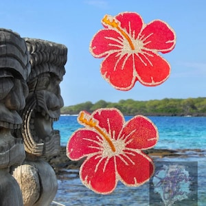 May include: Two red hibiscus flower decorations with gold glitter outlines and yellow stamens. The flowers are set against a backdrop of a tropical beach scene with wooden tiki statues and blue water.