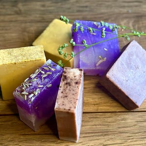 May include: A selection of handmade soaps in shades of yellow, purple, and brown. Some soaps are decorated with lavender and other plant-based elements. The soaps are displayed on a wooden surface, with a sprig of greenery for a natural aesthetic.