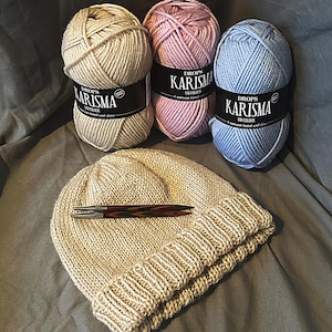 May include: A beige knitted beanie with a folded brim, next to three balls of yarn in beige, pink, and blue. Each yarn ball has a black label with white text that reads "Drops Karisma". A brown and black pen rests on the beanie.
