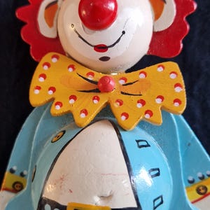 Könnte beinhalten: Holzclown-Ornament mit weißem Gesicht, roter Nase und rotem Haar. Der Clown trägt eine gelbe Fliege mit roten Punkten, eine blaue Jacke und eine gestreifte Hose. Das Ornament ist etwa 13 cm hoch.